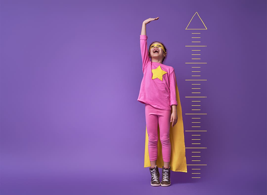 young girl in superhero costume stretching her arm above her head to measure her growth against a ruler on a bright purple wall