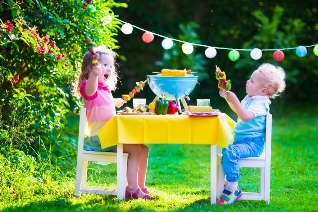 two toddlers eating food on skewers while sitting at a table set up outdoors on the grass
