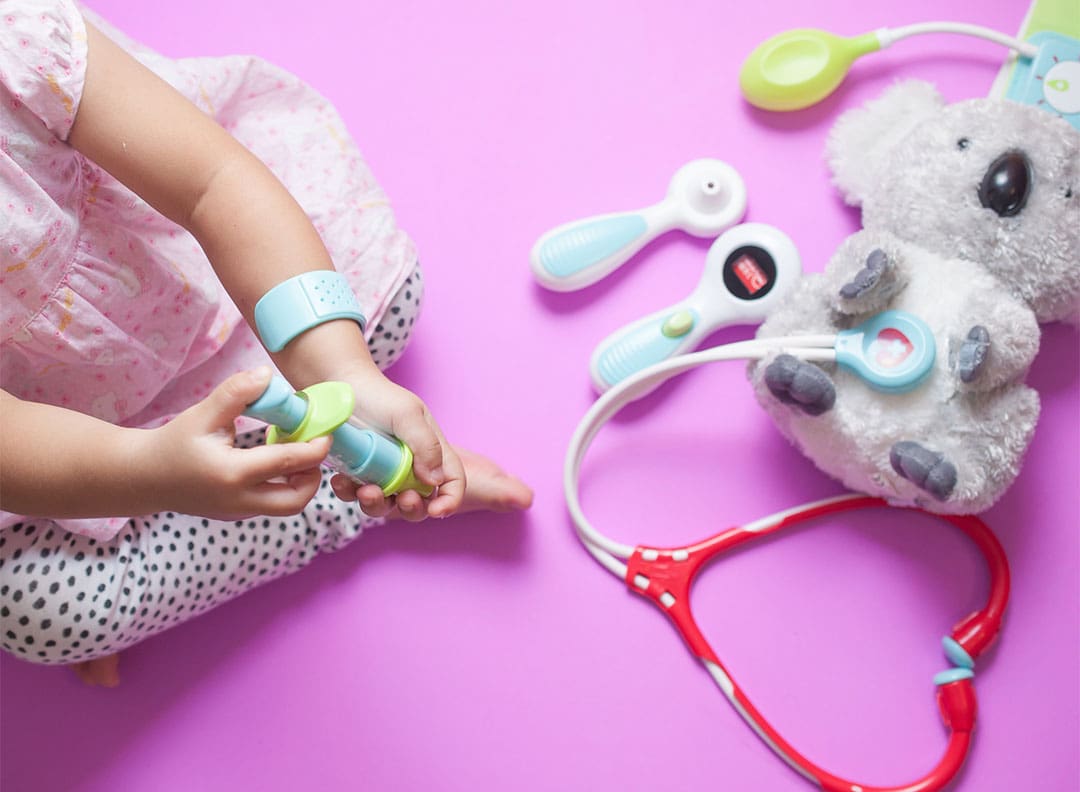 young child playing with a koala bear stuffie and toy doctor's equipment including plastic stethoscope, blood pressure cuff, and syringe